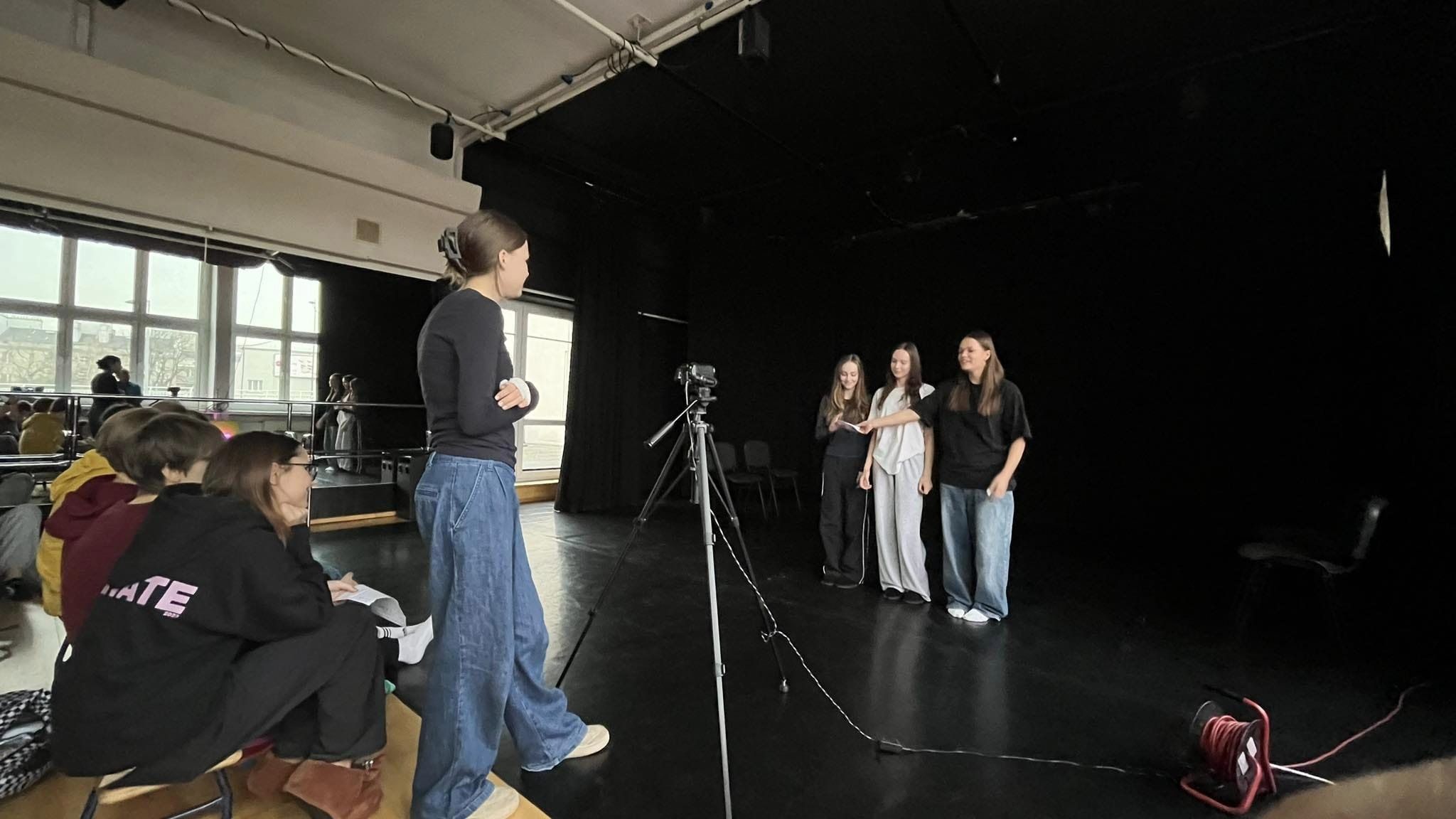 Trzy nastolatki stojące na scenie przed kamerą. Za kamerą stoi dziewczyna ubrana w czarną bluzkę i jeansy. Za nią na podłodze siedzi grupa osób obserwując scenę.