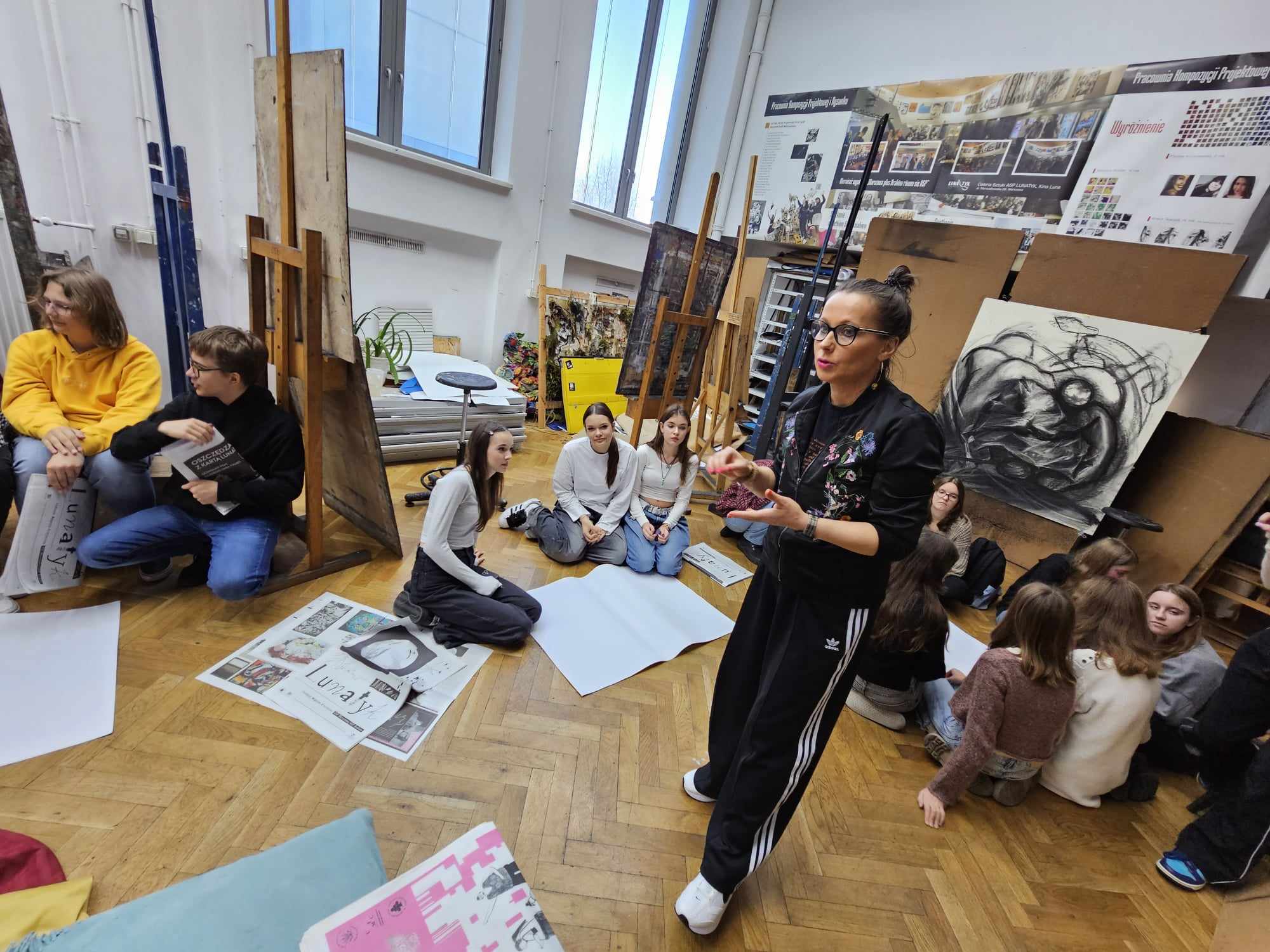 Grupa dzieci siedząca na podłodze z arkuszami papieru i przyborami do malowania. Wszyscy patrzą na kobietę stojącą między nimi 