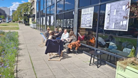 Grupa osób siedzi na krzesłach i leżakach przed oszklonym budynkiem z napisem "spokój", na budynku umieszczona wystawa, w tle chodnik , zieleń i budynki miejskie