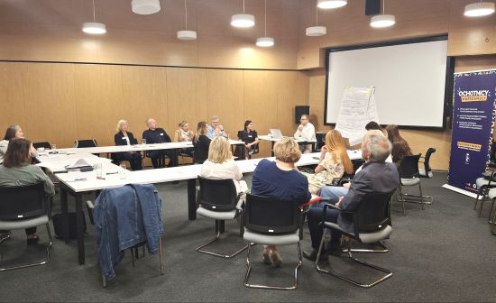 Grupa kilkunastu osób siedzących przy stole ustawionym w podkowę, w sali konferencyjnej