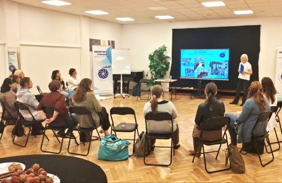 Zdjęcie wykonane w sali, kilkunastu uczestników siedzi na krzesłach ustawionych w półkole, słuchając prezentacji prowadzącej, na środku sali wyświetlona z rzutnika prezentacji i prowadząca stojąca frontem do uczestników