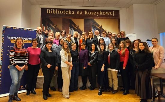 Zdjęcie zbiorowe: grupa ponad 30 osób stoi frontem do nas w budynku biblioteki, powyżej stojących osób napis: Biblioteka na Koszykowej, po lewej stronie granatowy roll-up z nazwą projektu "Ochotnicy warszawscy" 